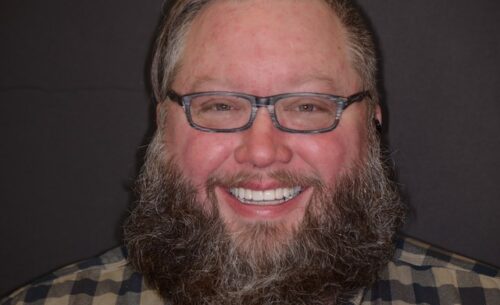Close-up of a smiling man with glasses and a thick beard wearing a plaid shirt against a dark background.