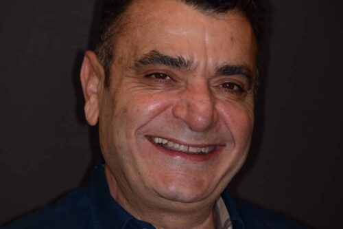 Close-up of a man smiling warmly, showing teeth, against a dark background.
