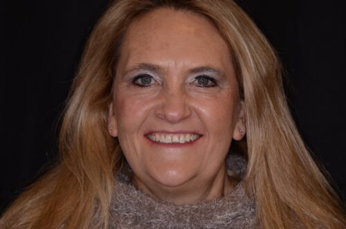 Smiling middle-aged woman with long blonde hair and blue eyes, wearing a gray knit turtleneck against a dark background
