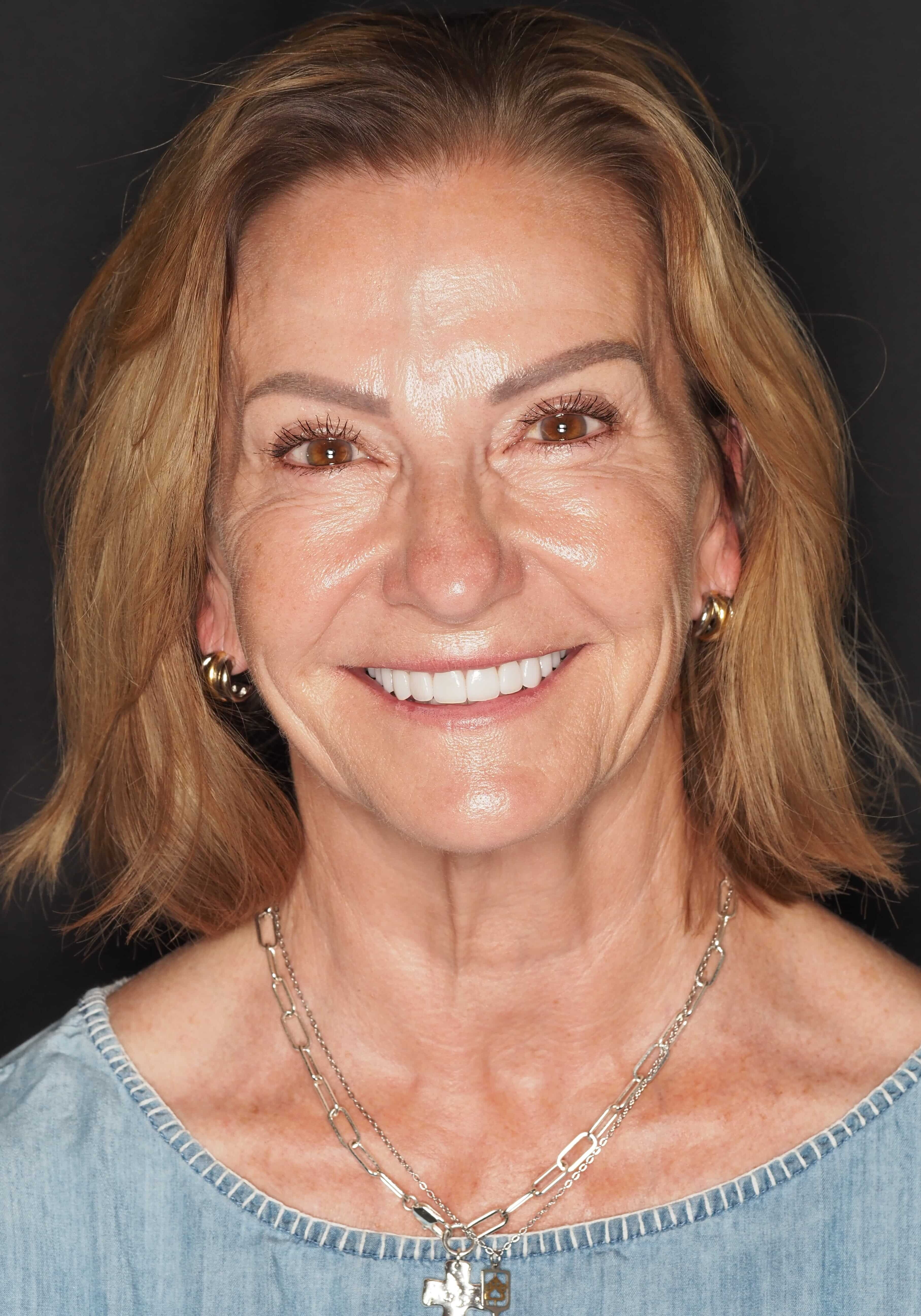 Smiling woman with light brown hair wearing a denim top and two silver necklaces, including a cross pendant, against a dark background.